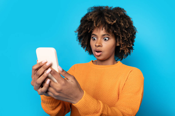 Schockiert schaut eine hübsche junge schwarze Frau mit buschigen Haaren in Orange auf den Smartphone-Bildschirm und grimassiert vor blauem Studiohintergrund, checkt spannende Geschäfte, bietet - Foto, Bild