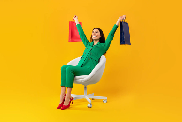 Plan complet du corps d'une femme accro au shopping satisfaite assise sur une chaise et levant les mains avec des sacs à provisions isolés sur fond de studio jaune, espace libre - Photo, image