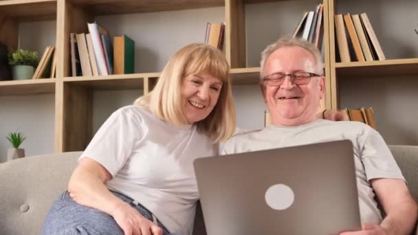 Vrolijke grootouders zitten op de bank, ze knuffelen en kijken naar het laptopscherm. Gepensioneerden met een Europees uiterlijk rusten - Video