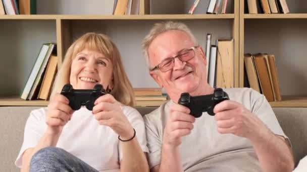 Joyeux couple retraité jouant à des jeux avec joystick. Un couple de personnes âgées, fasciné par un jeu de course automobile, est assis sur le canapé dans leur maison devant un écran de télévision. Bonne vie des retraités - Séquence, vidéo