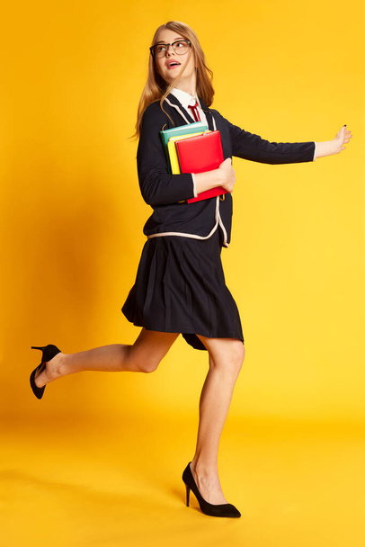 Running to lectures. Portrait of beautiful young girl, student in uniform with books over yellow studio background. Concept of youth, beauty, fashion, lifestyle, emotions, facial expression. Ad - Zdjęcie, obraz