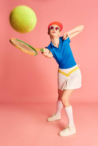 Hitting ball. Portrait of beautiful young girl in sportive clothes posing with tennis racket over pink background. Concept of youth, sport, fashion, lifestyle, emotions, facial expression. Ad - Photo, Image