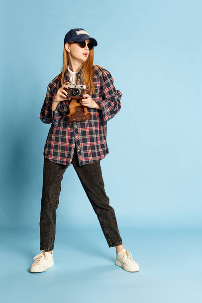 Traveller. Young beautiful girl in checkered shirt, cap and sunglasses posing with photo camera on blue studio background. Concept of youth, beauty, fashion, lifestyle, emotions, facial expression. Ad - Foto, afbeelding