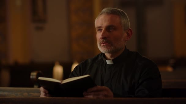 Een grijs-harige katholieke priester zit op een bank in een kerk met een bijbel in zijn handen. Een close-up van een katholieke priester die de bijbel leest. Hoge kwaliteit 4k beeldmateriaal - Video