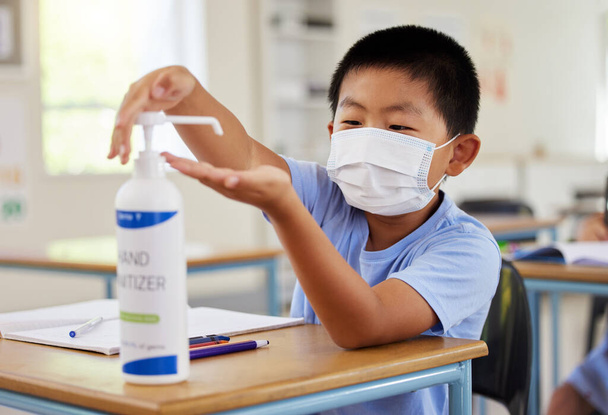 Covid, hand sanitizer and school student with mask cleaning, protecting and staying safe in education classroom. Small, little and cute asian boy in learning in study class preventing spread of virus. - Photo, Image
