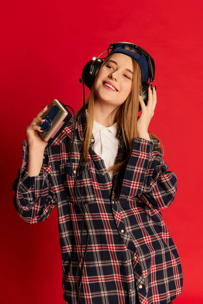 Young beautiful positive girl in checkered shirt and cap listening to music in headphones with player over red studio background. Concept of youth, beauty, lifestyle, emotions, facial expression. Ad - Foto, Imagen