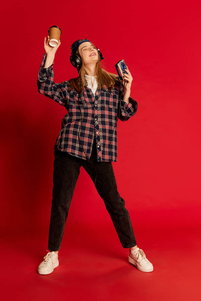 Energy. Young beautiful positive girl in checkered shirt and cap listening to music in headphones, dancing over red studio background. Concept of youth, fashion, lifestyle, emotions, facial expression - Фото, изображение