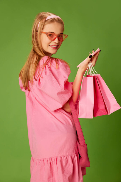 Beautiful blonde young girl in cute pink dress and trendy sunglasses posing with shopping bags on green studio background. Concept of youth, beauty, fashion, lifestyle, emotions, facial expression. Ad - Foto, imagen