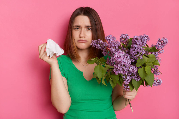 Ill girl with bouquet of lilac flowers, suffers from allergic malaise and rhinitis, seasonal disease, tired of sneezing, allergy to flowering holds handkerchief, poses isolated on pink wall background - Foto, imagen