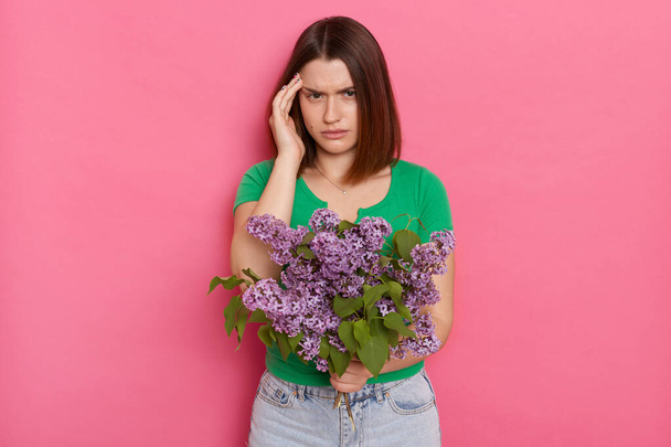 Portrait of sick young girl with brown hair wearing green casual t shirt holding bouquet of lilac flowers, suffering headache, posing isolated over pink wall background - Photo, image