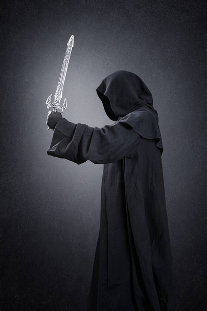 Dark knight with hooded cape and medieval sword over dark misty background - 写真・画像