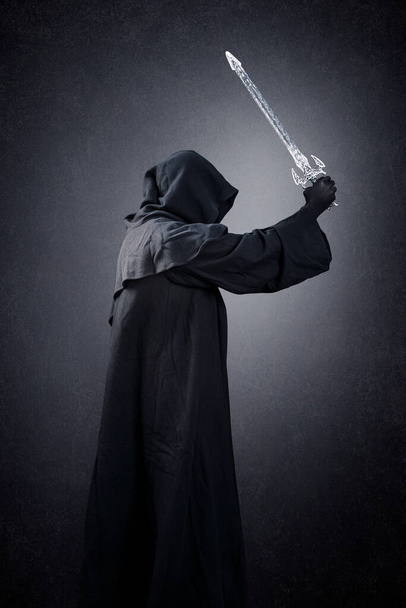 Dark knight with hooded cape and medieval sword over dark misty background - Фото, изображение