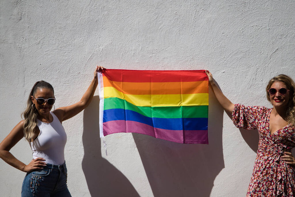 Két csinos fiatal nő napszemüvegben tartja a meleg büszkeség zászlaját a kezében. A nők boldogan ünneplik a meleg büszkeség napját. Egyenlőség fogalma, homoszexualitás, lgtb - Fotó, kép