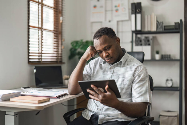 Черный бизнесмен напряжен, ему скучно и он слишком много думает, работая над планшетом в современном офисе. - Фото, изображение