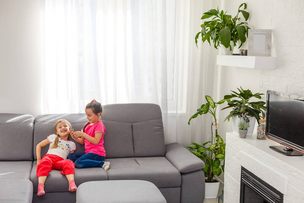 Залишайтеся вдома в безпеці. Двоє щасливих дітей сидять на затишному кріслі, розслабляючись, граючи у білій вітальні в приміщенні. Сестри розважаються вдома
 - Фото, зображення