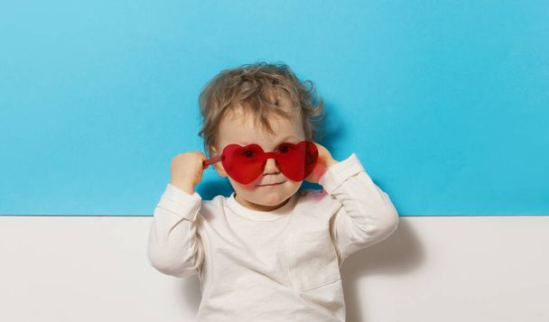 Portrait of surprised cute little baby boy in the heart shape sunglasses. Child having fun over blue background. Looking at camera. Wow funny face  - Фото, зображення