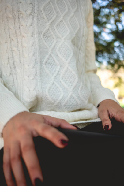 Κοντινό πλάνο του κοριτσιού με όμορφα χέρια, σκούρα νύχια και λευκό πουλόβερ (με λεπτομέρειες από ύφασμα) στο ηλιόλουστο πάρκο - Φωτογραφία, εικόνα