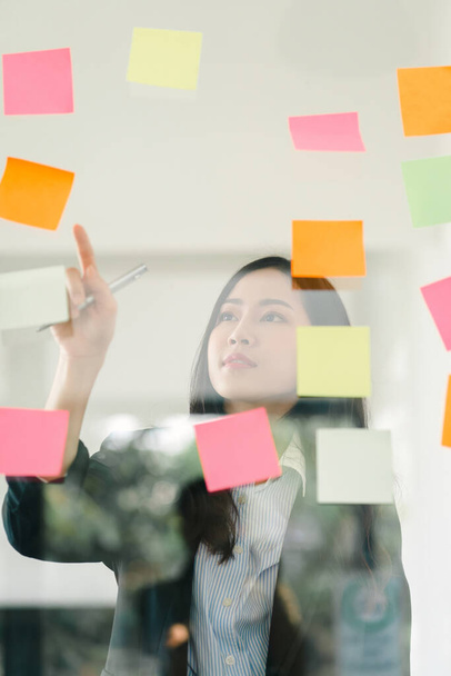 The concept of brainstorming is exemplified as a businesswoman shares ideas using Post-it notes, which she places on a glass wall - Foto, afbeelding