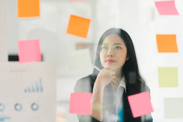 O conceito de brainstorming é exemplificado como uma mulher de negócios compartilha ideias usando notas Post-it, que ela coloca em uma parede de vidro - Foto, Imagem