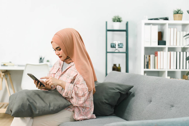 Portret azjatyckiej muzułmanki noszącej hidżab, korzystającej ze smartfona w domu, wysyłającej wiadomości lub przeglądającej portale społecznościowe siedząc na kanapie w salonie - Zdjęcie, obraz