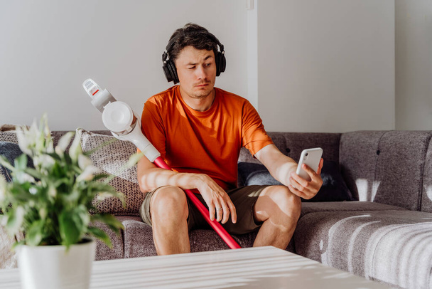 Man with wireless vacuum cleaner and headphones sitting on sofa checking messages in cellphone. White caucasian male tired of cleaning texting in smartphone holding vacuum cleaner wearing earphones. - Fotografie, Obrázek