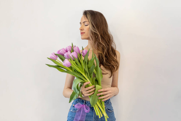 Slim young woman with a lilac bouquet of tulips on a white background. Standing with her eyes closed, wearing fashionable clothes - Photo, Image