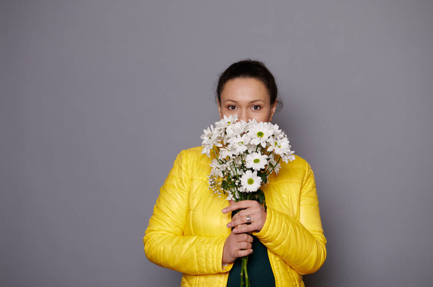 Portrait of a charming dark-haired woman with a bouquet of white spring flowers, looking confidently at camera, isolated over gray background. Beautiful women. People. Lifestyle. Festive event. - Fotó, kép