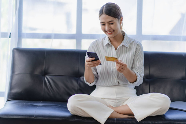 Азійка сидить на дивані і платить в інтернеті за смартфоном і кредитною карткою. - Фото, зображення