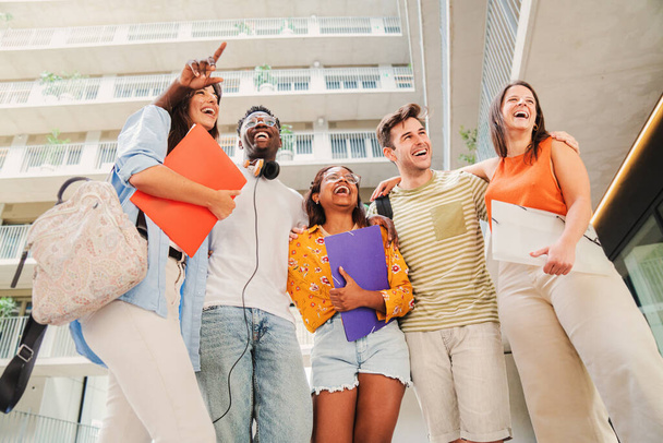 Eine Gruppe von multirassischen Teenagerstudenten lächelt und amüsiert sich gemeinsam auf dem Universitätscampus. Glückliche junge Leute stehen mit Rucksäcken und Notizbüchern am College. Freundschaftskonzept. Hoch - Foto, Bild