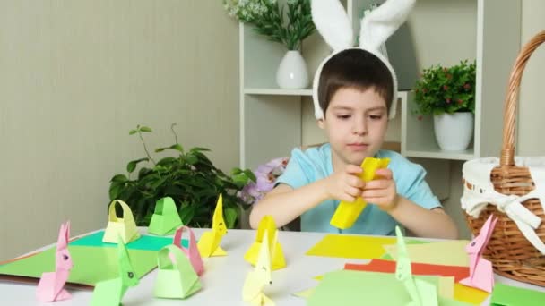 Menino pré-escolar com orelhas de coelho cabeça de nag faz artesanato de Páscoa, figurinhas de origami de coelhos e cestas de papel. - Filmagem, Vídeo