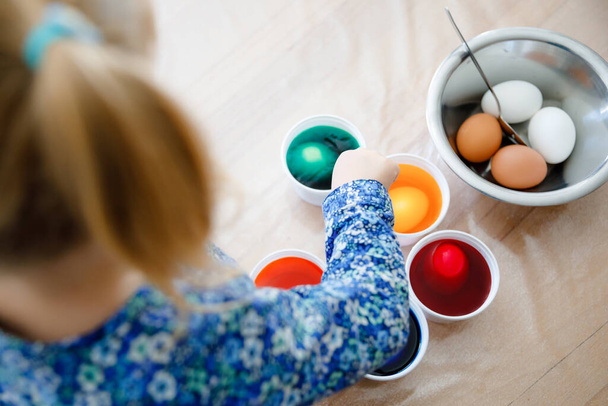 Closeup of little toddler girl coloring eggs for Easter. Close-up of child looking surprised at colorful colored eggs, celebrating holiday with family. From above, unrecognized face - Photo, image