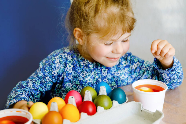 Aufgeregte kleine Mädchen färben Eier für Ostern. Kind schaut überrascht auf bunte Eier horten und feiert katholische und christliche Feiertage mit Familie. Nettes Kind hilft beim Färben, drinnen - Foto, Bild