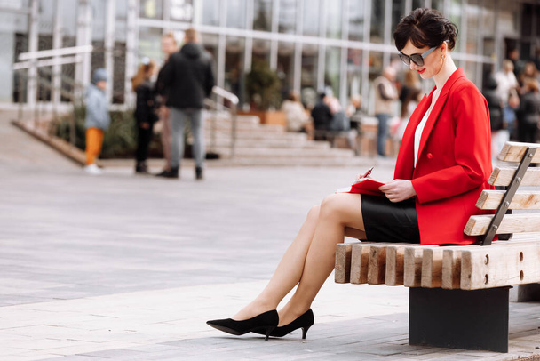 Business, paperityöt ja ihmiset käsite luottavainen liiketoiminnan nainen lukeminen ja kirjoittaminen muistiinpanoja muistiossa istuu kaupungin penkillä. Ajattelevainen nainen yllään punainen bleiseri ja nahka hame innolla. - Valokuva, kuva