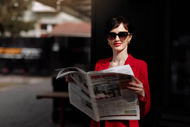 Стильная красивая брюнетка в красной куртке держит газету и читает новости по утрам на свежем воздухе возле офисного центра. Элегантная деловая женщина сделала перерыв на получение новой информации. - Фото, изображение