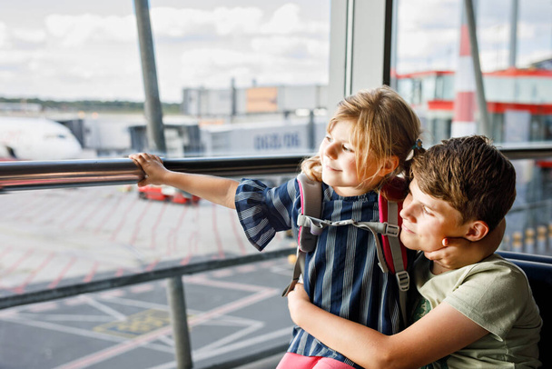 Little girl and school boy at the airport waiting for boarding at big window. Two kids stands at window against the backdrop of airplanes. Happy children, siblings leaving for family summer vacation. - Photo, Image
