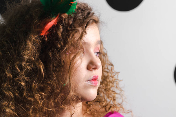 Πορτρέτο ενός γοητευτικού μικρού Curly-haired κοριτσιού, στολισμένο με πολύχρωμα φτερά, φοράει ένα ροζ φόρεμα ποζάροντας σε ένα ασπρόμαυρο πουά δωμάτιο. - Φωτογραφία, εικόνα