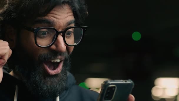 Amazed happy bearded Indian businessman in glasses receive sms message read good news with smartphone at parking lot. Excited male winner Arabian man celebrating success win victory with mobile phone - Footage, Video