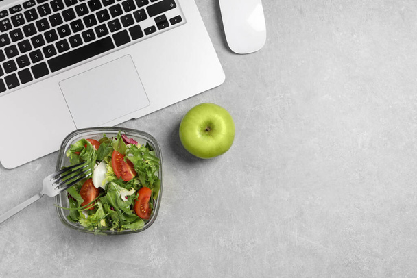 Fresh vegetable salad, apple, fork and laptop on light grey table at workplace, flat lay. Business lunch - Photo, image