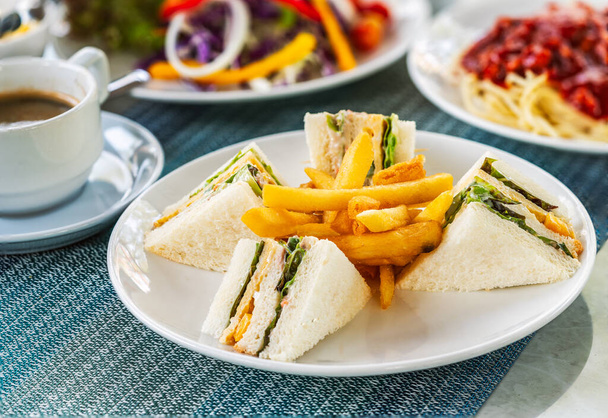 Ontbijt, vier sandwichclubs en samen gebakken op een wit bord op een tafel, wazige achtergrond van ander voedsel, salade en koffiekop. - Foto, afbeelding