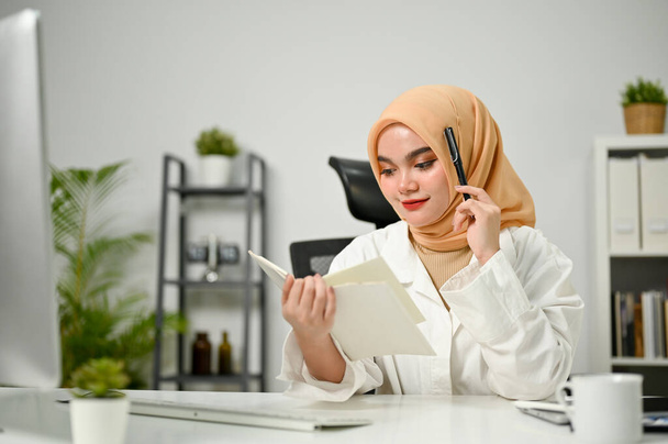 Attraente e professionale donna d'affari musulmana asiatica millenaria che indossa hijab leggendo alcune informazioni su un libro alla sua scrivania in ufficio. - Foto, immagini