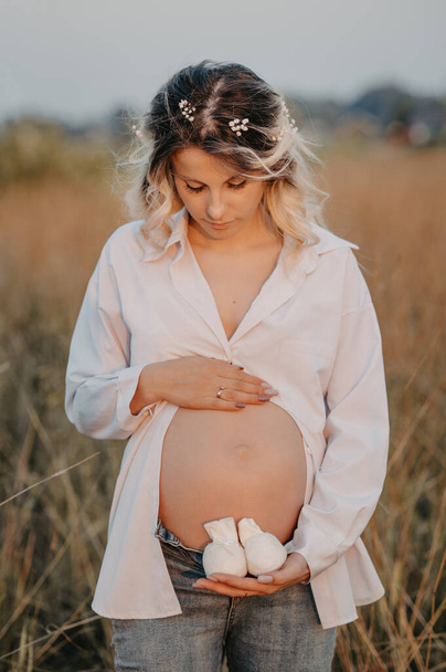 A young expectant mother in the third trimester of pregnancy in a white dress hugs her stomach and holds booties against the background of the natural landscape. The concept of future motherhood - Photo, Image