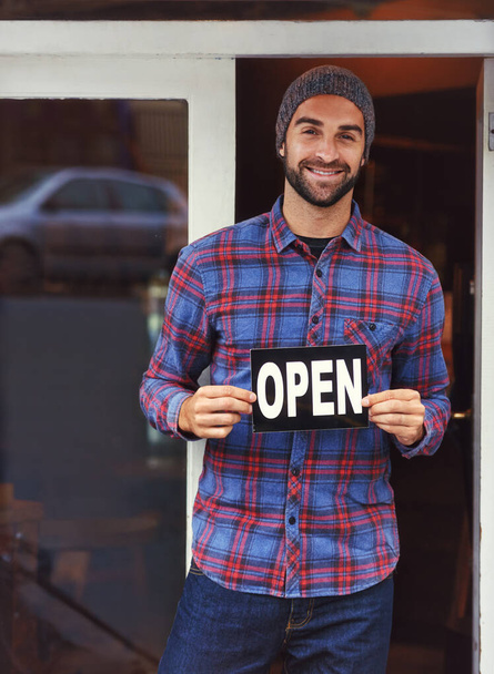 Ven a echar un vistazo dentro. un joven guapo parado junto a su negocio y sosteniendo el letrero abierto - Foto, imagen