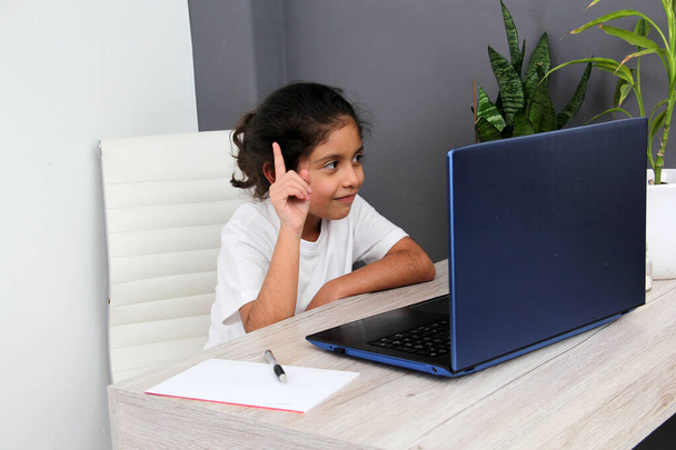 9-year-old κορίτσι Latino κάνει κατ 'οίκον εκπαίδευση παίρνει online μαθήματα στο σπίτι σε ένα γραφείο με ένα φορητό υπολογιστή, μελέτες, είναι έκπληκτος και συμμετέχει στην τάξη - Φωτογραφία, εικόνα