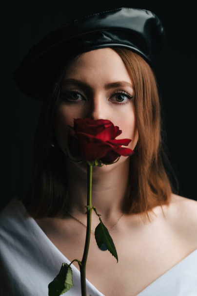 黒いベレー帽の美しい少女の肖像画。彼女の顔には赤いバラを持つパリの女性のイメージのスタジオで若い女の子。赤い唇 - 写真・画像
