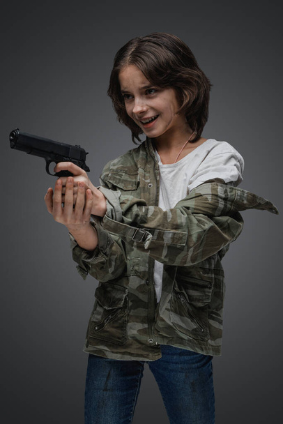 Shot of mad young girl with camouflage clothes with pistol against gray background. - Photo, image