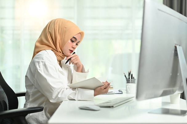 Enfocado millennial musulmana mujer de negocios con hijab sentado en su lugar de trabajo y comprobar el plan de trabajo. - Foto, Imagen