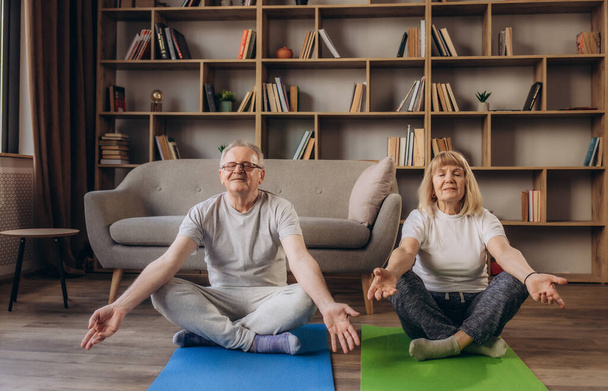 Smiling elderly husband and wife sit on floor at home meditate with mudra hands, happy overjoyed active senior spouses teach learn yoga practice in living room. Healthy lifestyle, stress free concept - Photo, Image