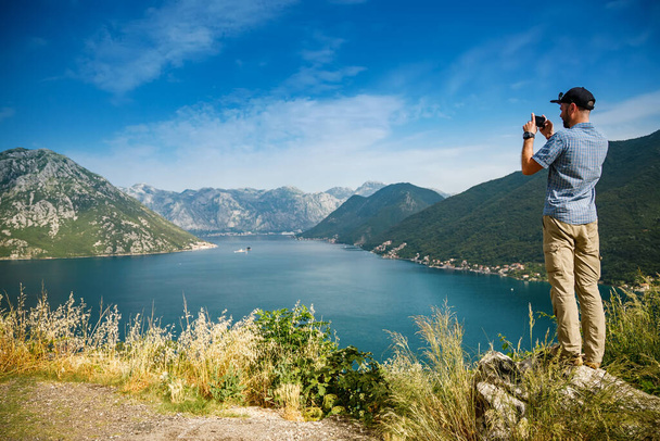 Tourist man taking a picture on his smartphone, standing on the edge of a cliff, enjoying amazing sea view of the Bay of Kotor, Montenegro - Foto, Imagen
