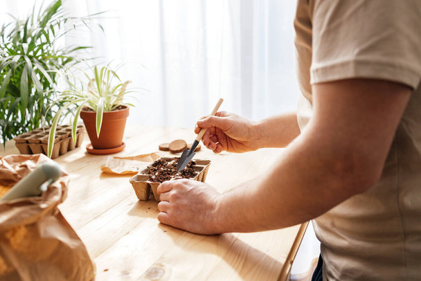 The process of planting seeds. A man pours earth with a small spatula into ecological peat moulds. Landscaping and gardening in the apartment.  - Photo, image