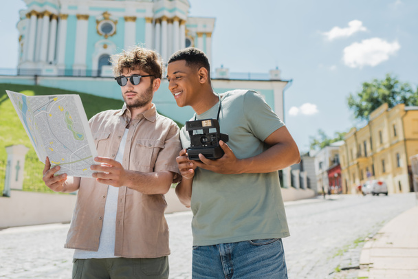 Genç tur rehberi, mutlu Afrika kökenli Amerikalı turistin yakınındaki şehir haritasına bakıyor. Andrews 'un Kyiv' e inişini gösteren eski model bir kamerayla.  - Fotoğraf, Görsel
